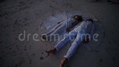 幸福的新婚夫妇在日落时躺在沙滩上，穿着新娘的婚纱。 拍摄的最初想法.. A.
